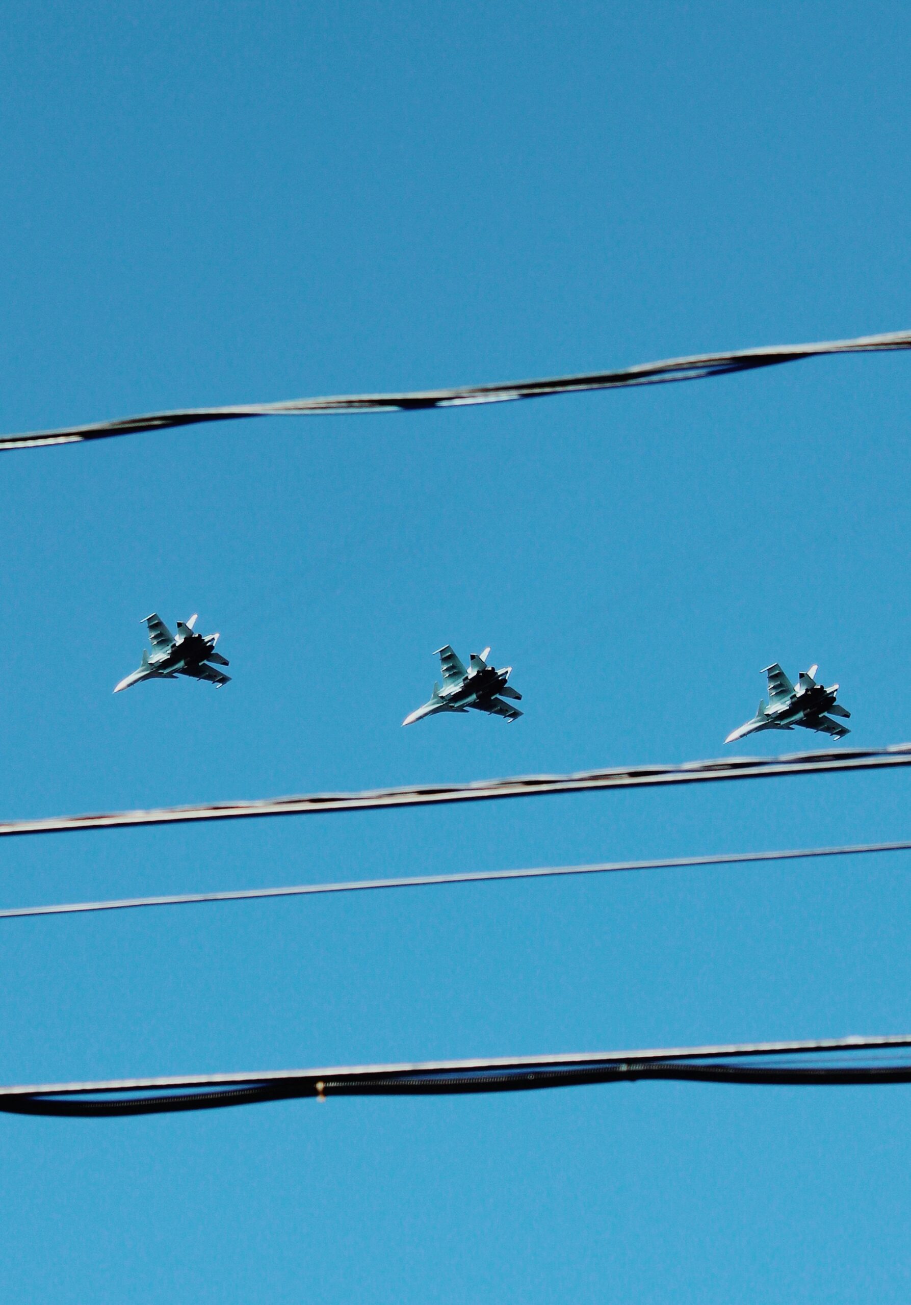 Three fighter jets soar in formation above electric wires on a clear day.